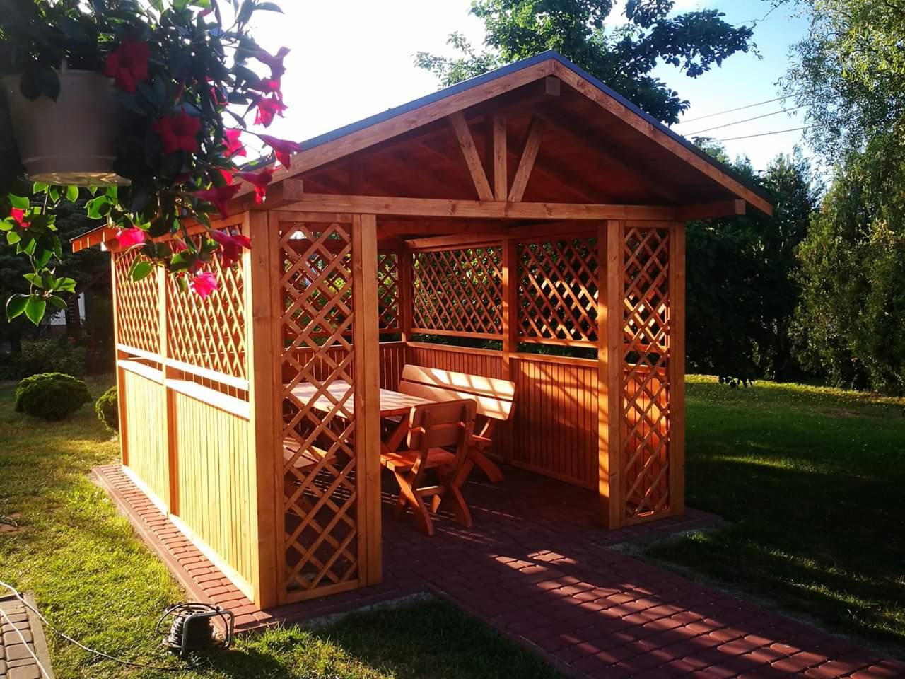 Drewniana altana ogrodowa z kratownicowymi ścianami, wyposażona w stół i ławki, otoczona zielonym trawnikiem i ścieżką z czerwonej kostki brukowej, z wiszącymi kwiatami w donicy w lewym górnym rogu.