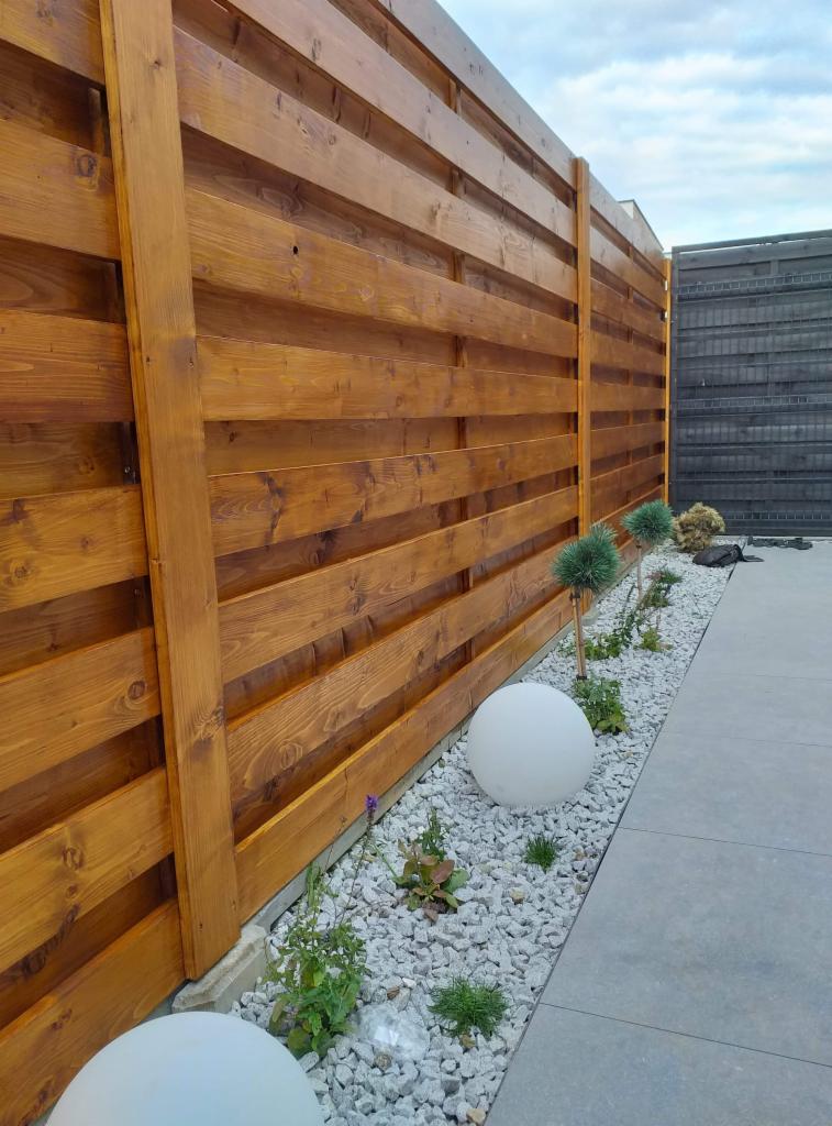 Nowoczesne ogrodzenie drewniane w Wieliczce, z poziomych desek o ciepłym, brązowym odcieniu, oddzielające ogród z białymi kulami-lampami i drobnymi roślinami od sąsiedniej posesji z ciemnym...