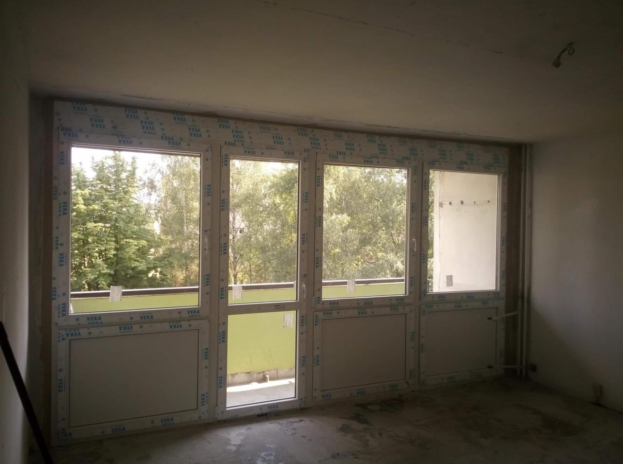 Wnętrze pokoju z zamontowanymi oknami balkonowymi z białymi ramami z widokiem na zieleń za oknem, widoczne niedokończone ściany i podłoga.