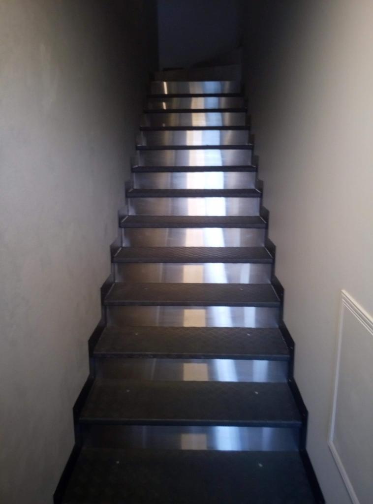 Schody wewnętrzne z metalowymi stopniami i ciemnymi krawędziami, prowadzące w górę w słabo oświetlonym korytarzu o szarych ścianach.