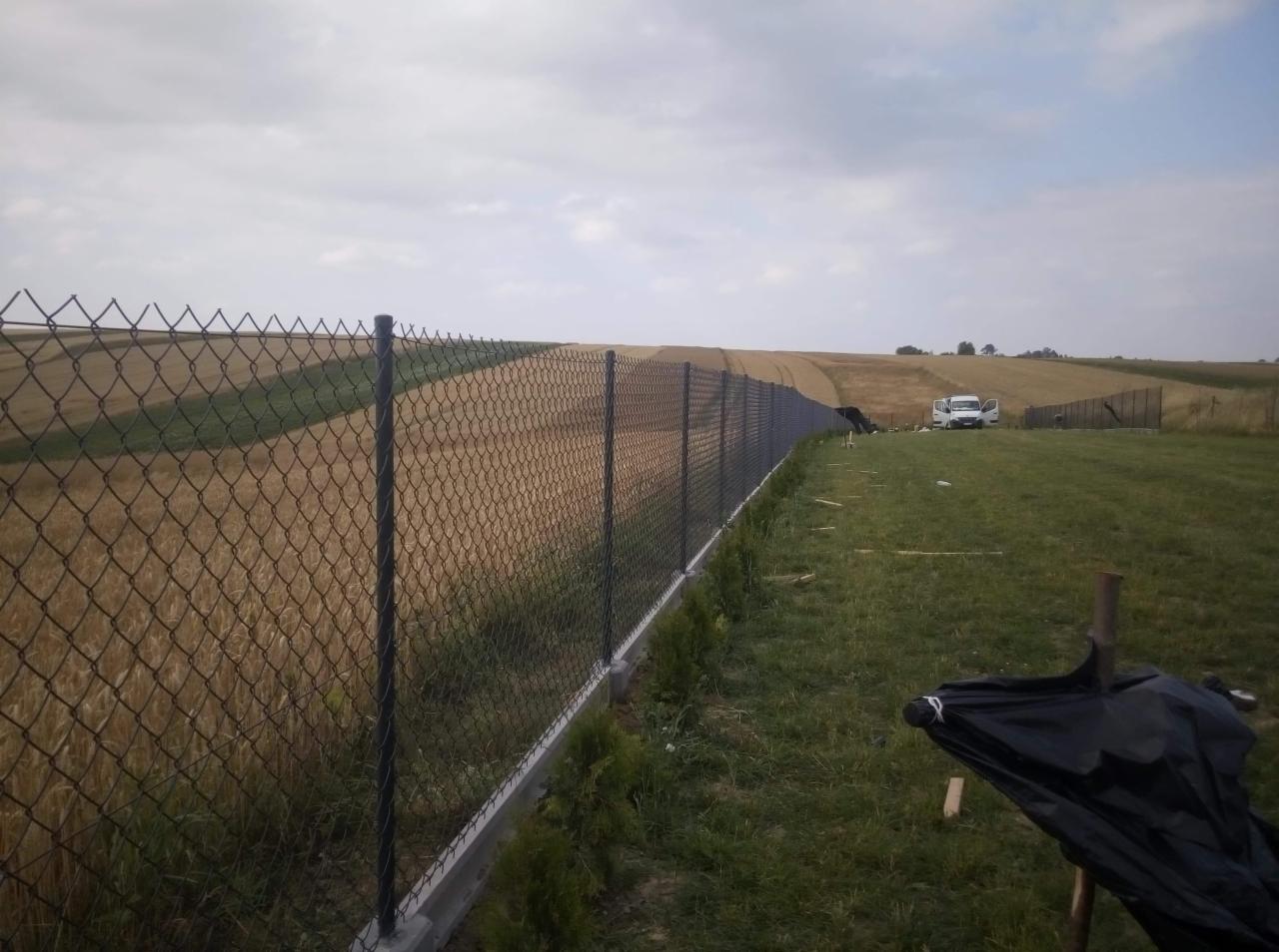 Nowo postawione ogrodzenie z siatki na betonowej podmurówce, oddzielające pole uprawne od trawnika, z widocznym samochodem dostawczym w tle.
