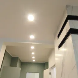 Równy rząd wpuszczanych lamp sufitowych w nowym korytarzu z zielonymi ścianami i kolumną wykończoną marmurem z czarnymi akcentami.