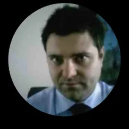 Portret mężczyzny w jasnoniebieskiej koszuli i ciemnym krawacie, umieszczony w okrągłej ramce na czarnym tle, z poważnym wyrazem twarzy.