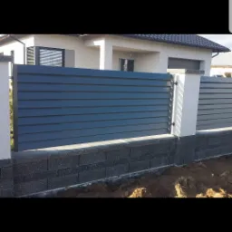 Nowoczesne ogrodzenie panelowe w kolorze grafitowym, z podmurówką z szarej kostki, na tle białego domu jednorodzinnego.