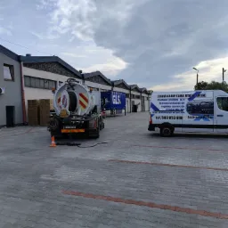 Specjalistyczny pojazd WUKO do udrażniania kanalizacji na brukowanym placu, z widocznym wężem w studzience. Obok zaparkowany bus z reklamą firmy.