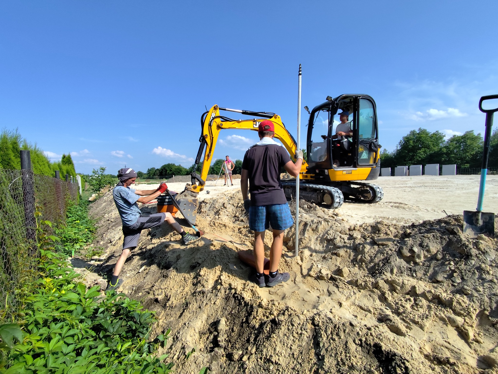 Żółta mini koparka w akcji na placu budowy, pracownicy mierzą poziom terenu, w tle widoczne płyty betonowe i ogrodzenie z siatki.