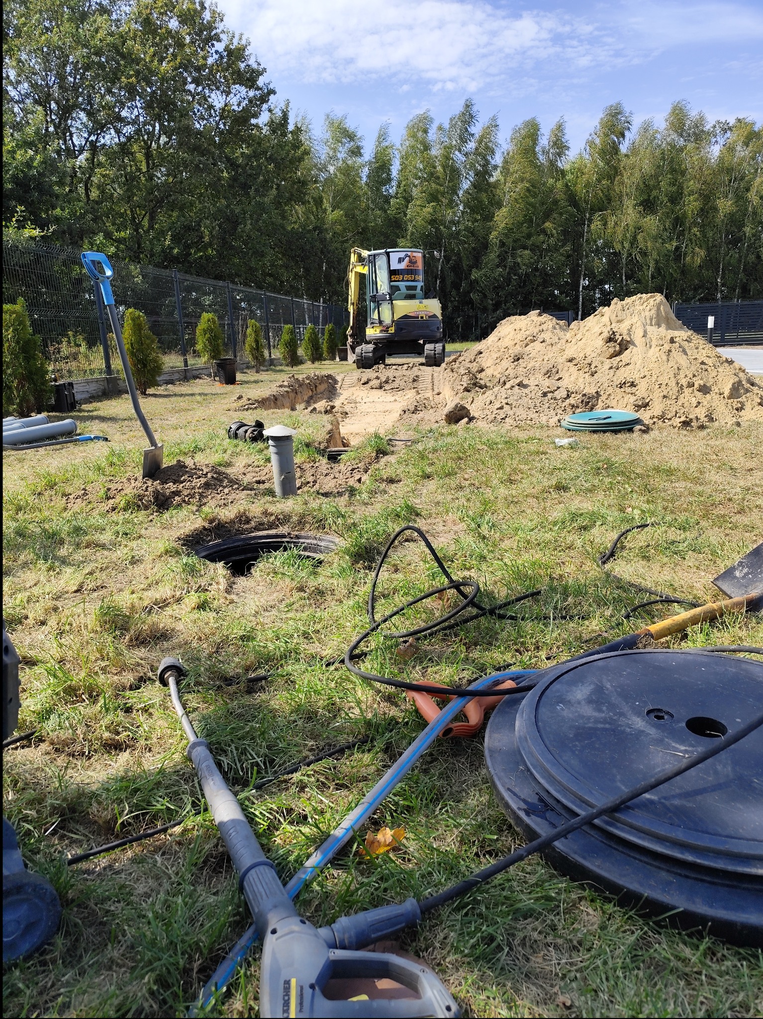 Wykonanie wykopu pod instalację sanitarną na trawiastym terenie, widoczny mini-koparka, rury, wykop z otworem, stos ziemi i narzędzia pracy, w tle drzewa i ogrodzenie.