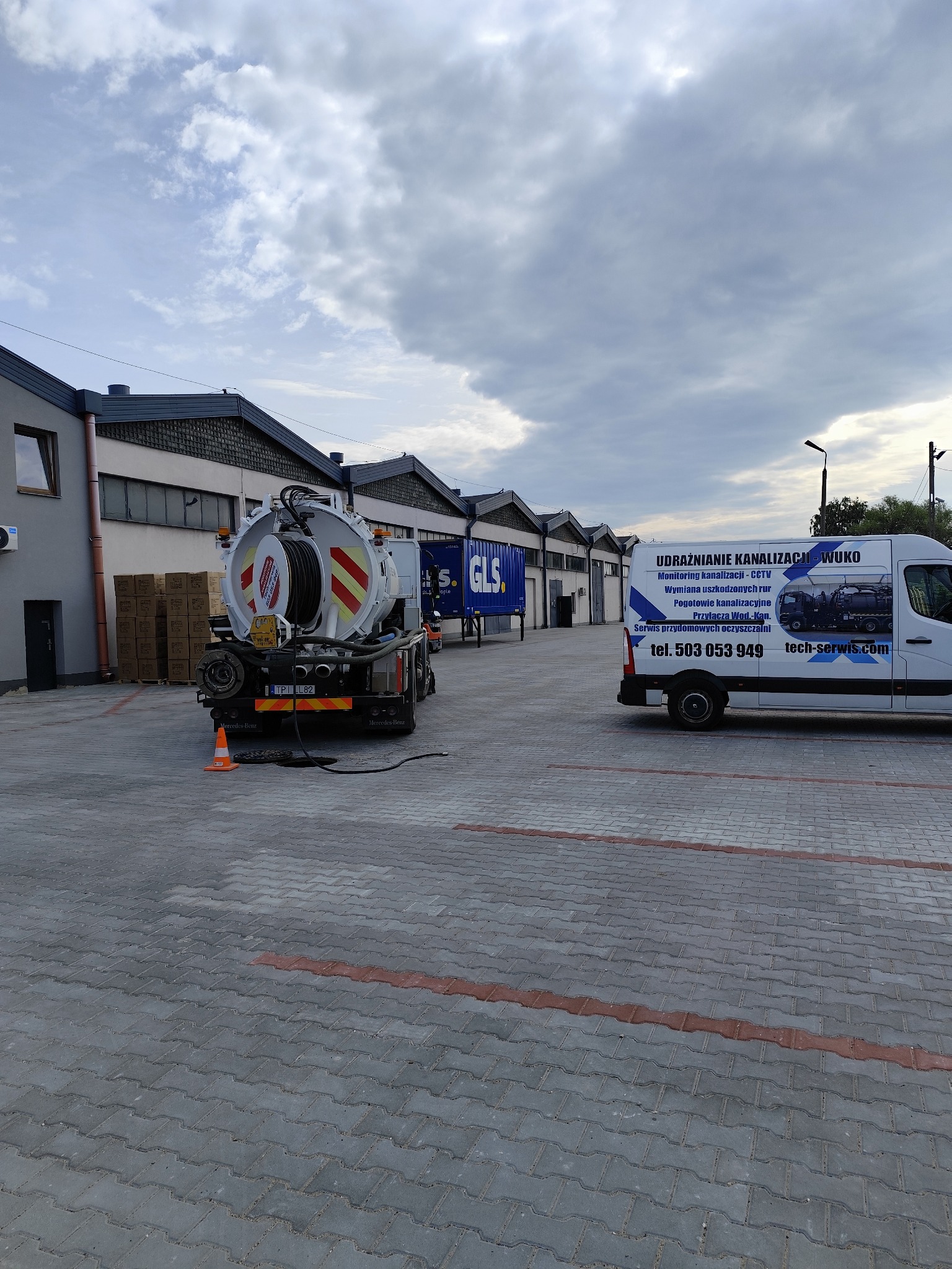 Specjalistyczny pojazd WUKO do udrażniania kanalizacji na brukowanym placu, z widocznym wężem w studzience. Obok zaparkowany bus z reklamą firmy.