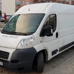 Biały samochód dostawczy Fiat Ducato zaparkowany na brukowanym parkingu przed blokiem mieszkalnym, widoczny numer rejestracyjny RT 45105.