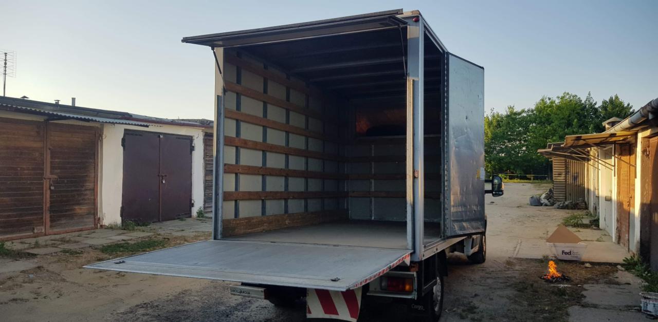 Otwarta pusta paka samochodu dostawczego z drewnianymi listwami zabezpieczającymi ściany, widoczna z zewnątrz na podwórku z garażami, obok palenisko z płomieniami i pojemnik FEDEX.