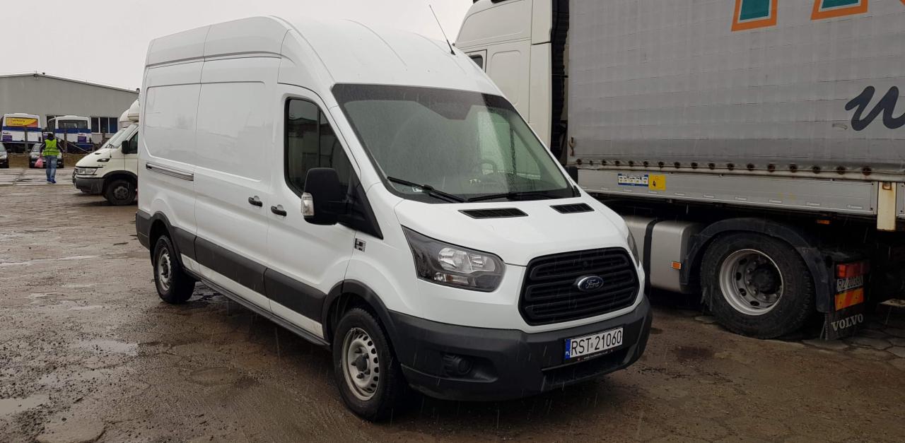 Biały bus Ford Transit z widoczną tablicą rejestracyjną RST 21060 stoi na mokrym placu, w tle zaparkowana naczepa z niebiesko-pomarańczowym logo.
