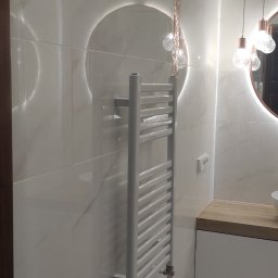 ArtHome - Nowoczesna łazienka z białymi marmurkowymi płytkami, podświetlanym okrągłym lustrem, grzejnikiem drabinkowym i drewnianym blatem umywalki.