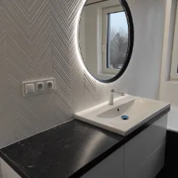 Nowoczesna łazienka z umywalką nablatową na szafce z czarnym blatem, okrągłe lustro z podświetleniem LED i białe płytki ułożone w jodełkę na ścianie.