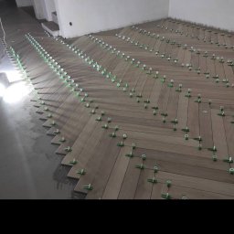 ArtHome - Układanie drewnopodobnych płytek podłogowych w jodełkę, z użyciem systemu poziomowania płytek (klipsy i kliny) na podłożu betonowym, widoczne instalacje elektryczne na ścianie.