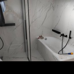 ArtHome - Wykończona łazienka z marmurowymi płytkami, czarną armaturą i szklaną kabiną prysznicową, widoczna wanna z akcesoriami do czyszczenia.