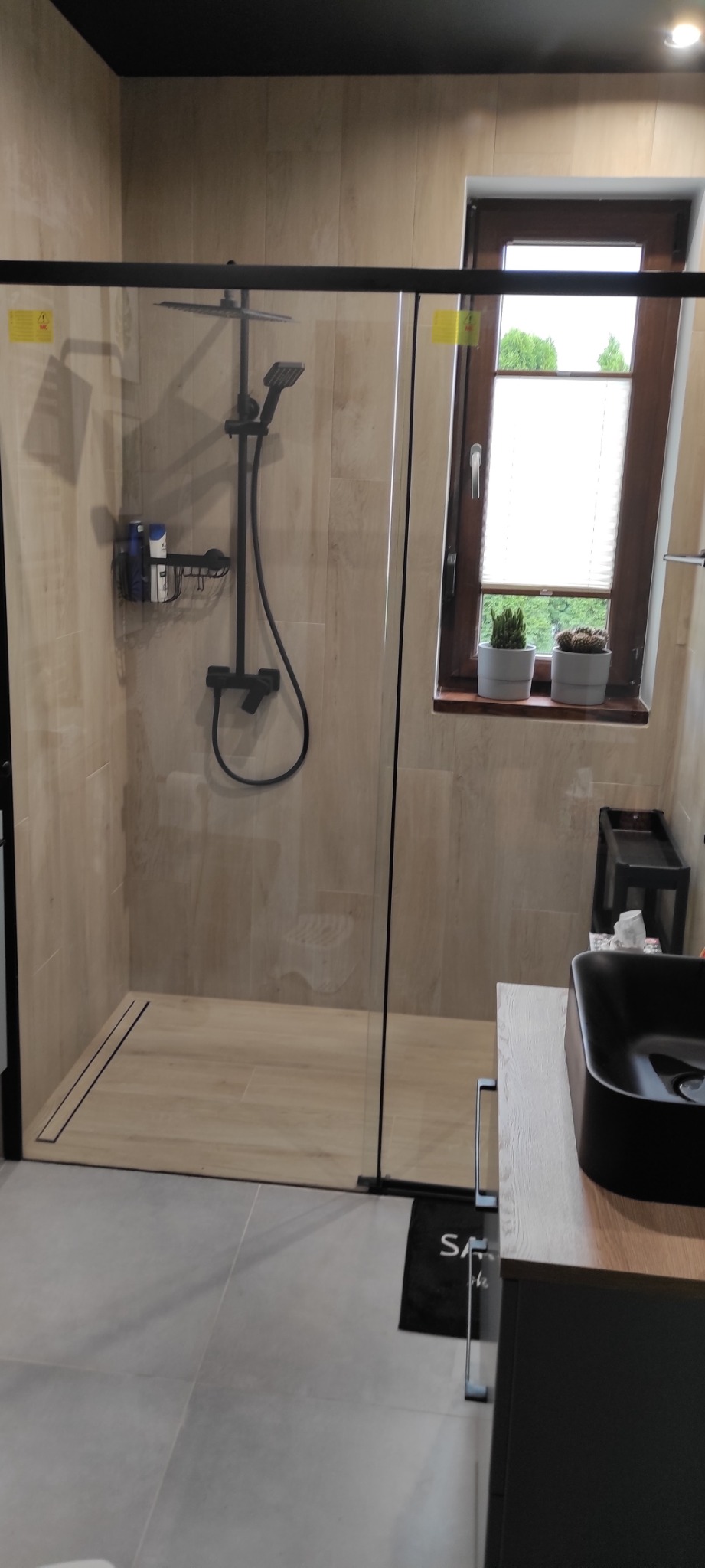 Nowoczesna łazienka z kabiną prysznicową typu walk-in, wykończona płytkami imitującymi drewno, z czarną armaturą i odpływem liniowym, oraz oknem z widokiem na zieleń.