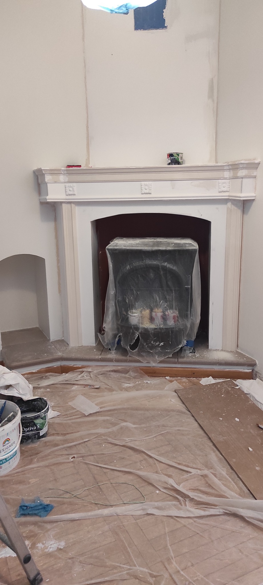 Przygotowania do malowania: kominek zabezpieczony folią, ściany po wstępnym malowaniu, widoczne puszki z farbą na podłodze przykrytej folią ochronną.