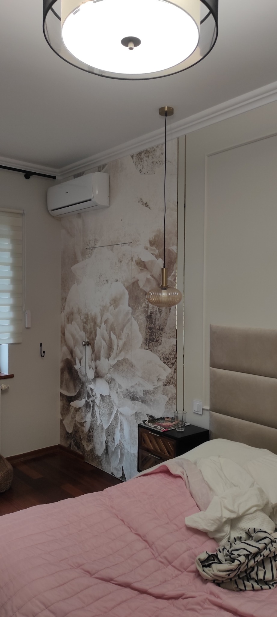 Sypialnia z łóżkiem w różowej pościeli, szafką nocną z lampką, klimatyzatorem i tapetą z motywem kwiatowym na ścianie.