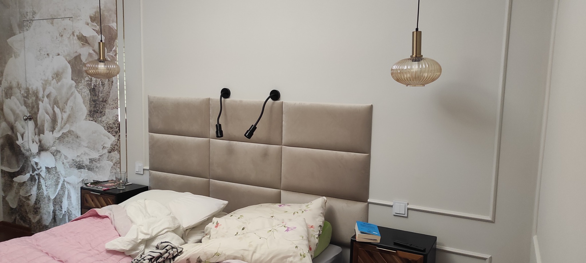 Sypialnia z beżowym panelem tapicerowanym za łóżkiem, lampkami nocnymi na giętkich ramionach, wiszącymi lampami z żłobionego szkła i szafkami nocnymi z elementami drewnianymi oraz fototapetą...