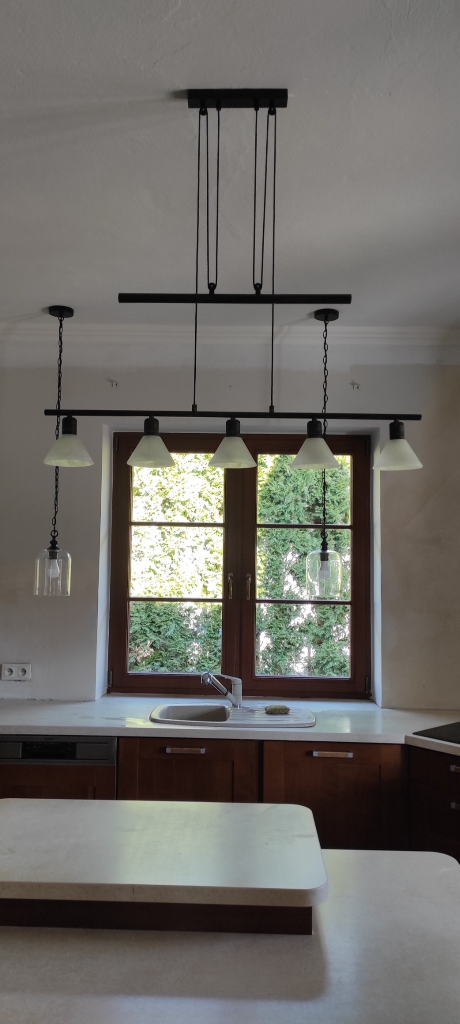 Nowoczesna kuchnia z brązowymi szafkami, marmurowym blatem i oknem z widokiem na zieleń, oświetlona czarną lampą sufitową z kilkoma kloszami.