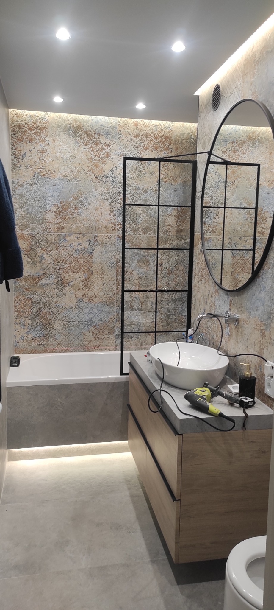 Nowoczesna łazienka z umywalką nablatową, kabiną prysznicową typu walk-in z czarnymi szprosami i okrągłym lustrem; na blacie leżą narzędzia i akcesoria łazienkowe.