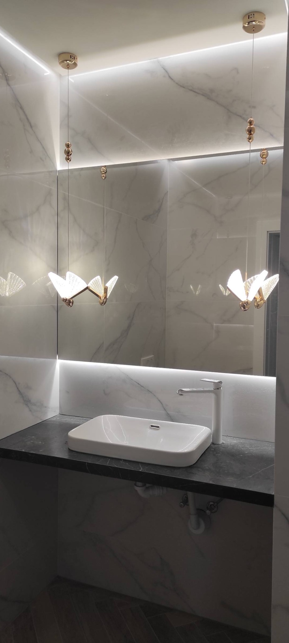 Elegancka łazienka z marmurowymi płytkami, białą umywalką nablatową na czarnym blacie, nowoczesną armaturą i dekoracyjnym oświetleniem w stylu motyla.