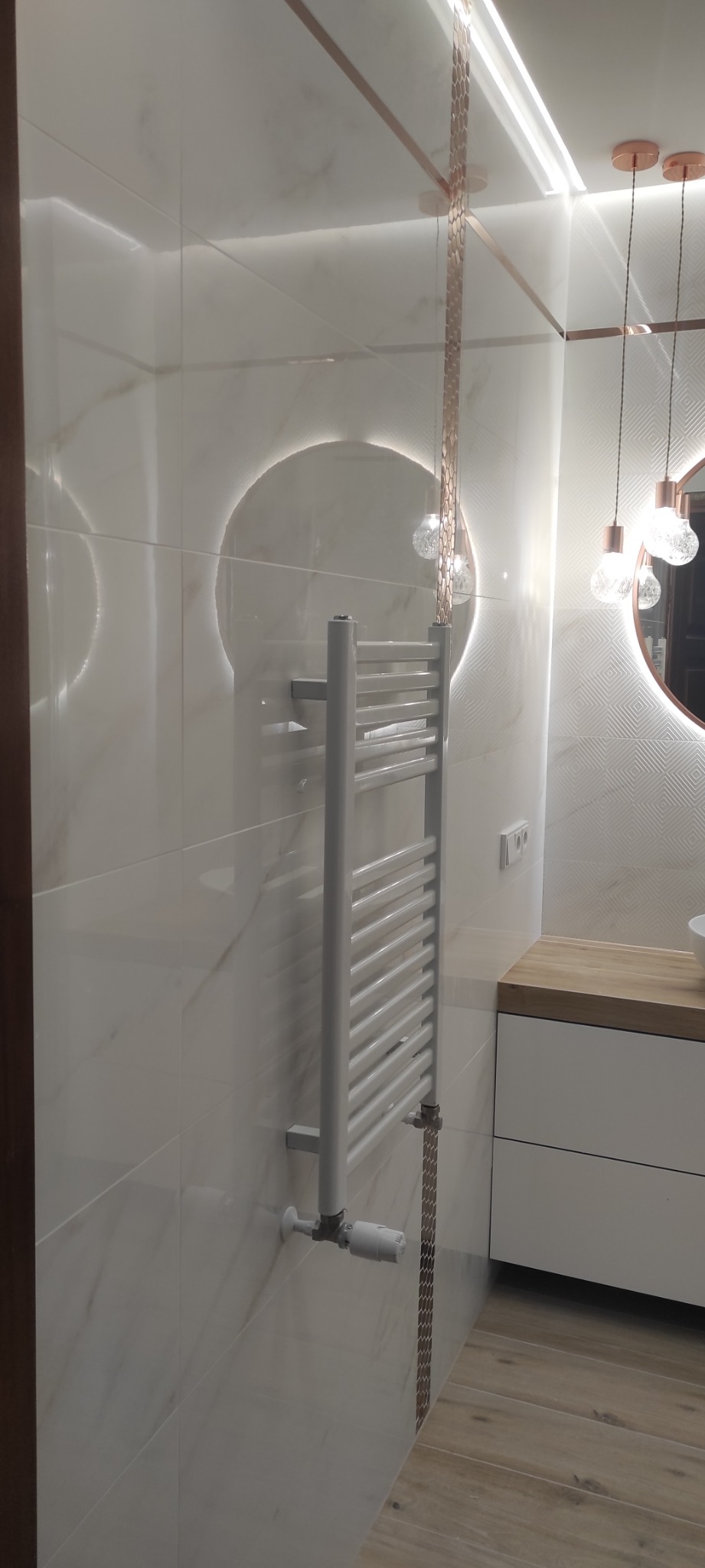 Nowoczesna łazienka z białymi marmurkowymi płytkami, podświetlanym okrągłym lustrem, grzejnikiem drabinkowym i drewnianym blatem umywalki.