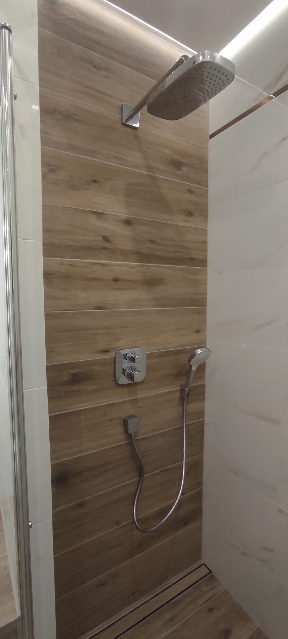 Nowoczesna kabina prysznicowa z drewnopodobnymi panelami ściennymi, chromowana armatura, deszczownica i szklane drzwi.