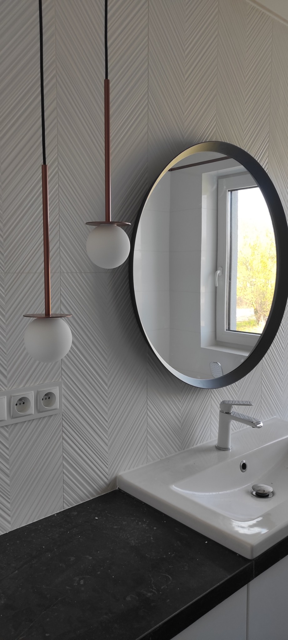 Nowoczesna łazienka z umywalką nablatową, okrągłym lustrem w czarnej ramie, dwoma wiszącymi lampami z kulistymi kloszami i białymi płytkami 3D w jodełkę na ścianie.