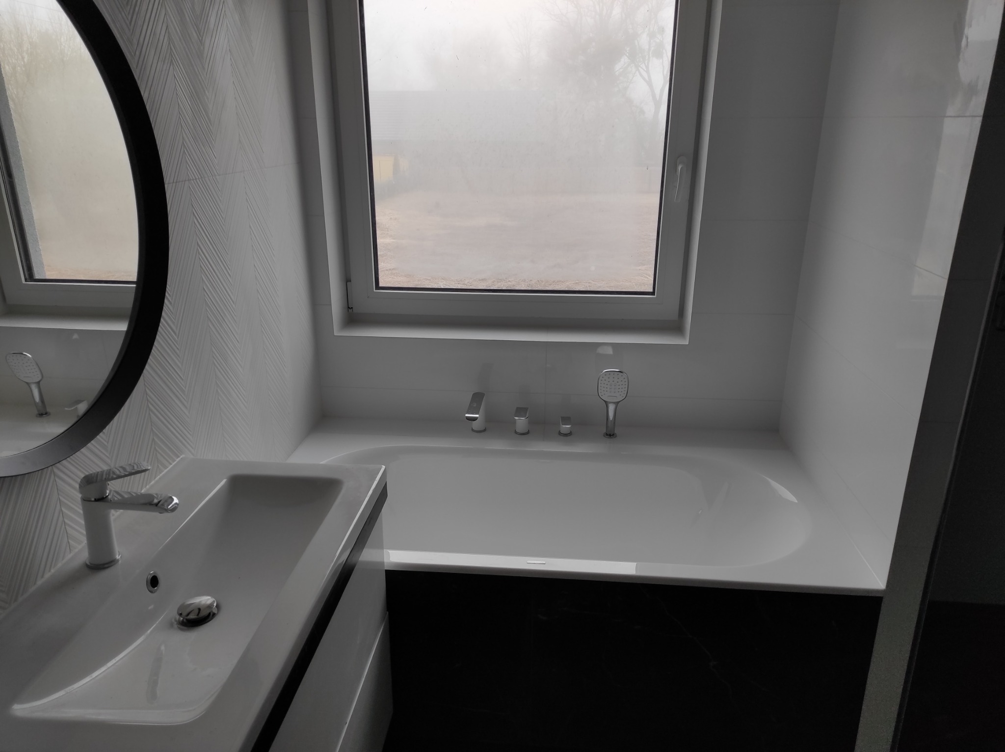 Nowoczesna łazienka z białą armaturą, umywalką z szafką i wanną pod oknem, wykończona płytkami w odcieniach szarości i czerni, z okrągłym lustrem.