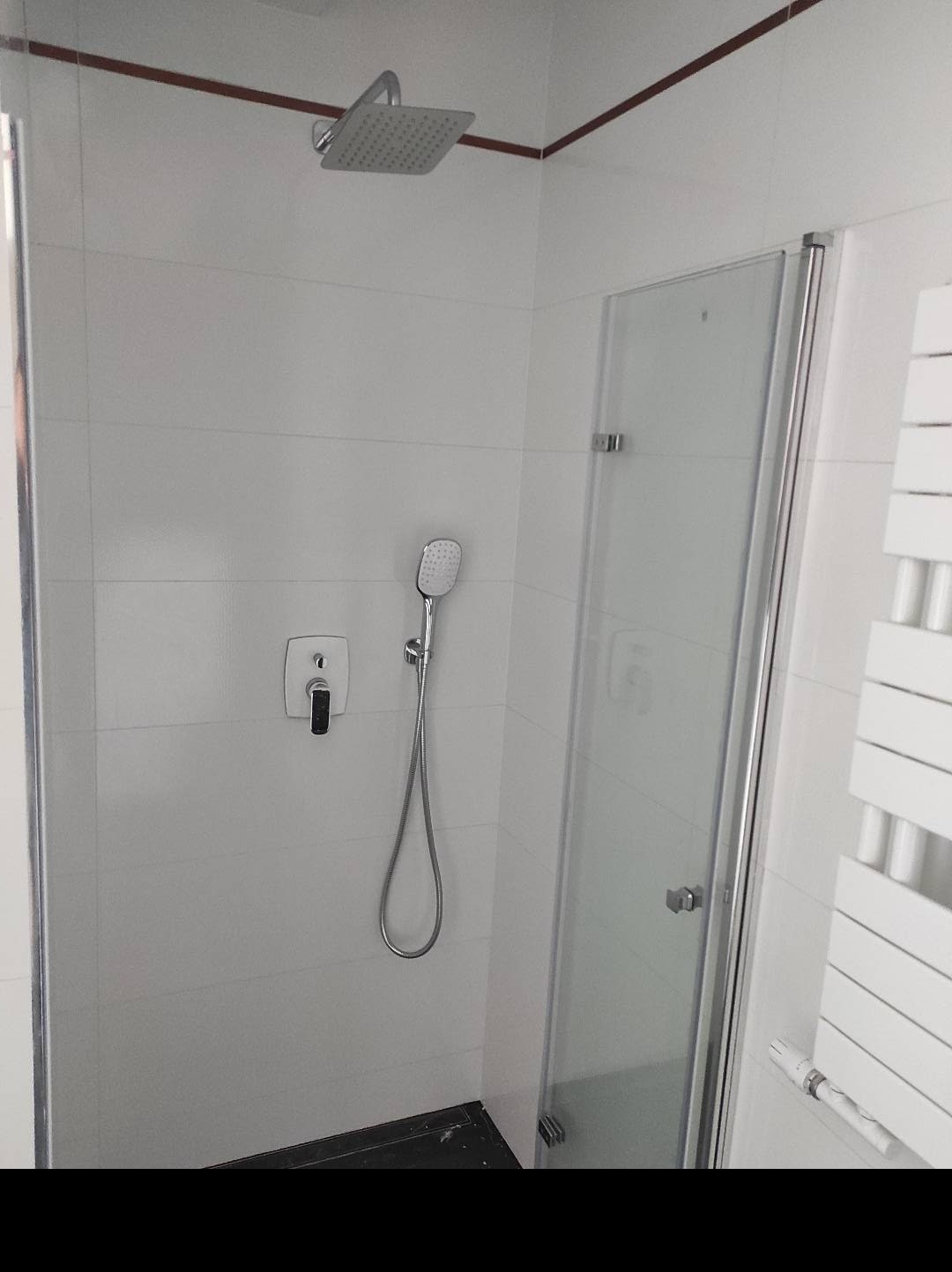 Nowoczesna kabina prysznicowa z deszczownicą i panelem natryskowym, wykończona białymi płytkami z ciemną fugą, widoczny grzejnik drabinkowy.