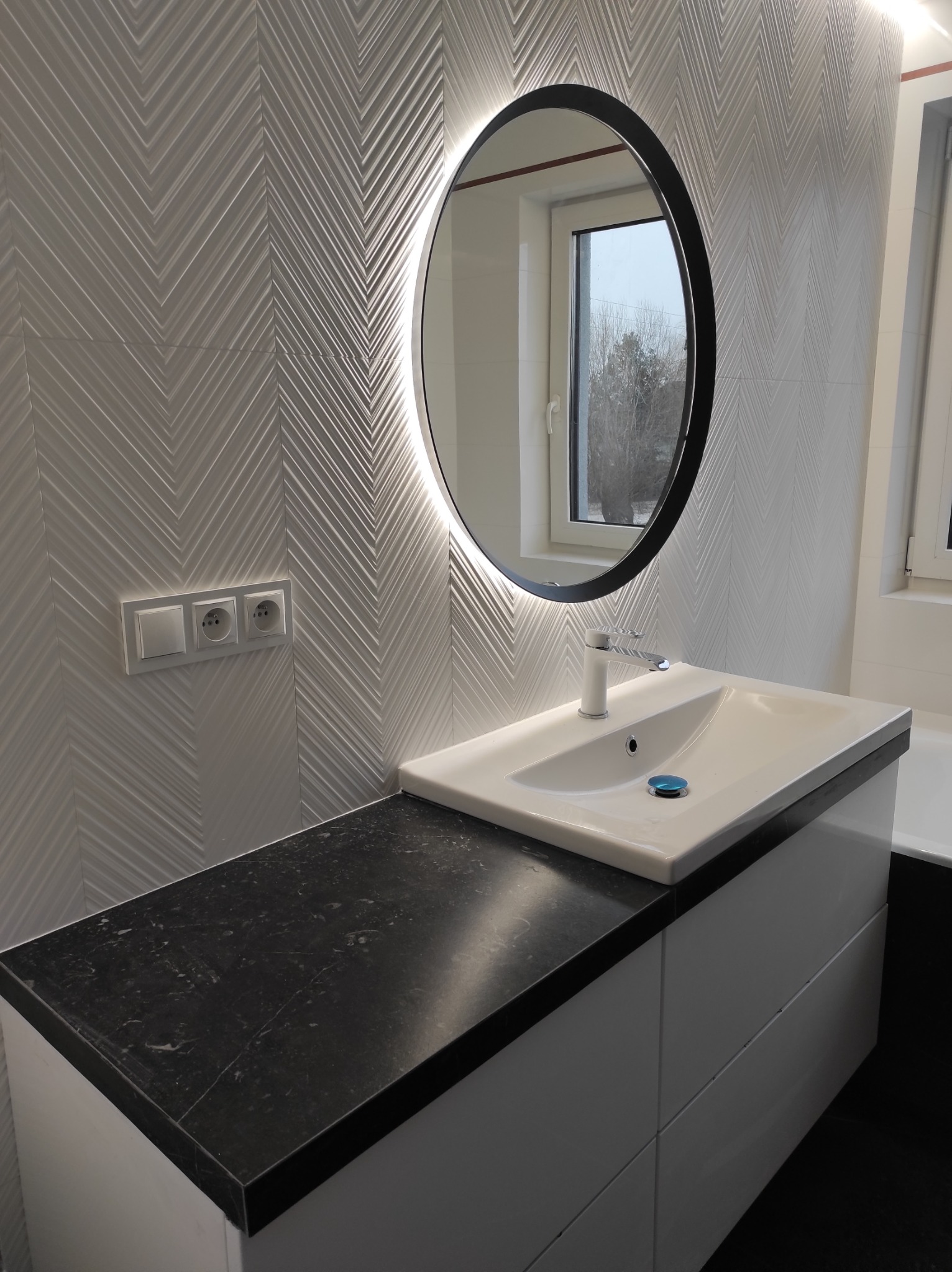 Nowoczesna łazienka z umywalką nablatową na szafce z czarnym blatem, okrągłe lustro z podświetleniem LED i białe płytki ułożone w jodełkę na ścianie.