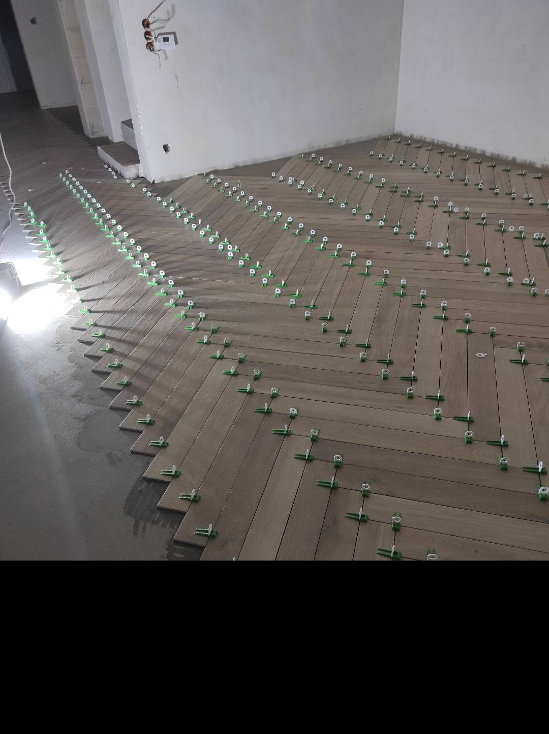 Układanie drewnopodobnych płytek podłogowych w jodełkę, z użyciem systemu poziomowania płytek (klipsy i kliny) na podłożu betonowym, widoczne instalacje elektryczne na ścianie.