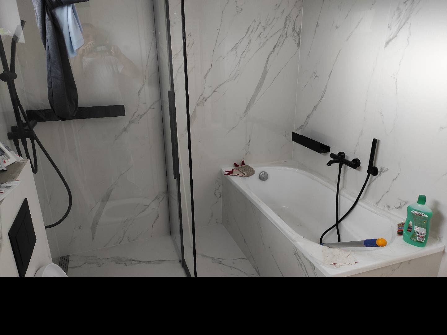 Wykończona łazienka z marmurowymi płytkami, czarną armaturą i szklaną kabiną prysznicową, widoczna wanna z akcesoriami do czyszczenia.