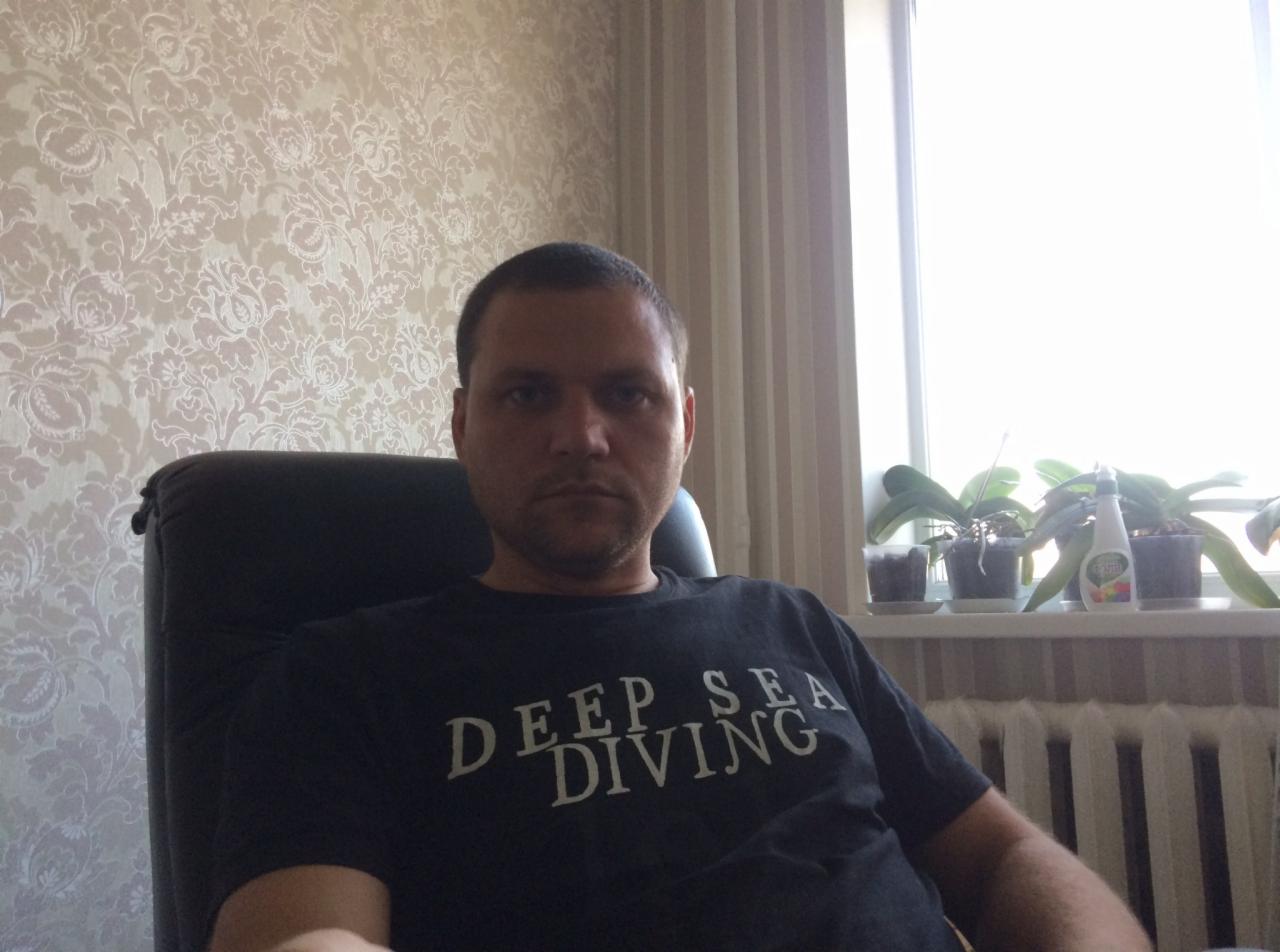 Mężczyzna siedzi na krześle na tle wzorzystej tapety i okna z roślinami, ubrany w czarną koszulkę z napisem 'DEEP SEA DIVING'.