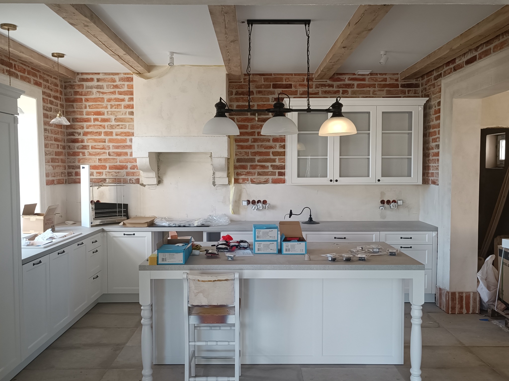 Kuchnia w trakcie wykańczania: białe szafki, ceglana ściana, drewniane belki stropowe, wyspa kuchenna z narzędziami i akcesoriami, surowa podłoga, widoczne ślady prac remontowych.