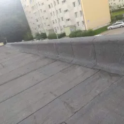 Płaski dach pokryty papą termozgrzewalną z widocznym obrzeżem attyki i blokami mieszkalnymi w tle.