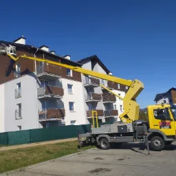 Żółty podnośnik koszowy na tle budynku wielorodzinnego z brązową elewacją, kosz podnośnika ustawiony przy dachu, błękitne niebo w tle.