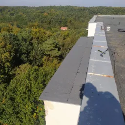 Widok z góry na płaski dach pokryty papą, z obróbką blacharską i narzędziem leżącym na niej, w tle las i wieża telekomunikacyjna.