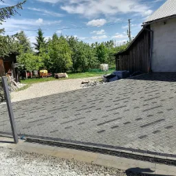 Ułożona kostka brukowa w odcieniach szarości z ciemniejszymi akcentami, widoczny system odprowadzania wody, na posesji z budynkami gospodarczymi i zielenią w tle.