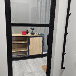 Loftowe drzwi wewnętrzne z czarną ramą i trzema szybami, widok na wnętrze z półkami i narzędziami, minimalistyczny design.