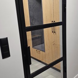 Syska Jakub Usługi Spawalnicze - Czarna, przeszklona ścianka działowa z widokiem na zabudowę meblową z siedziskiem. Minimalistyczny design, jasne drewno, szare siedzisko, nowoczesne wnętrze.