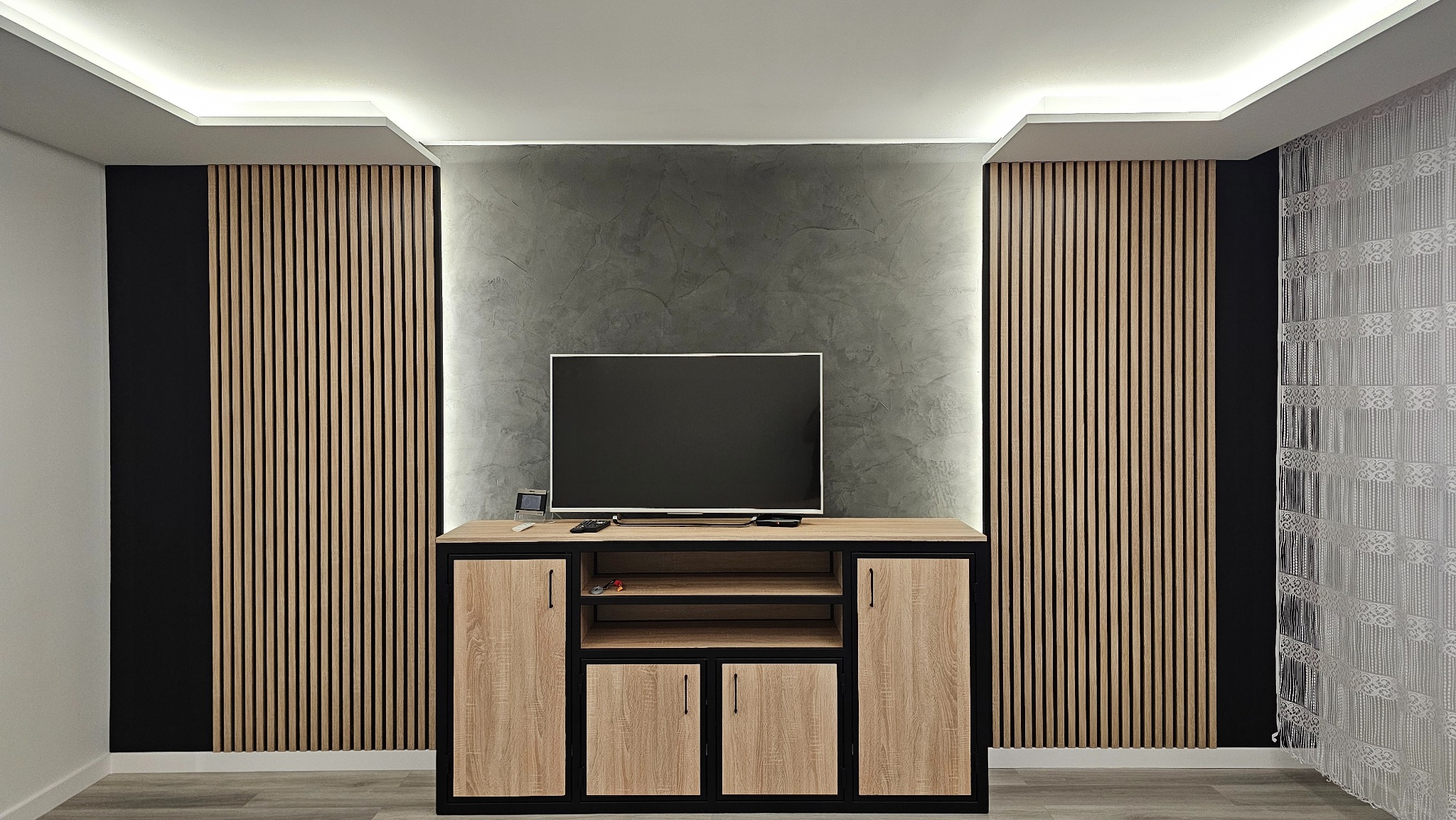 Salon z drewnianymi lamelami na ścianach, szarą ścianą i szafką RTV. Centralnie umieszczony telewizor. Oświetlenie LED podkreśla aranżację.