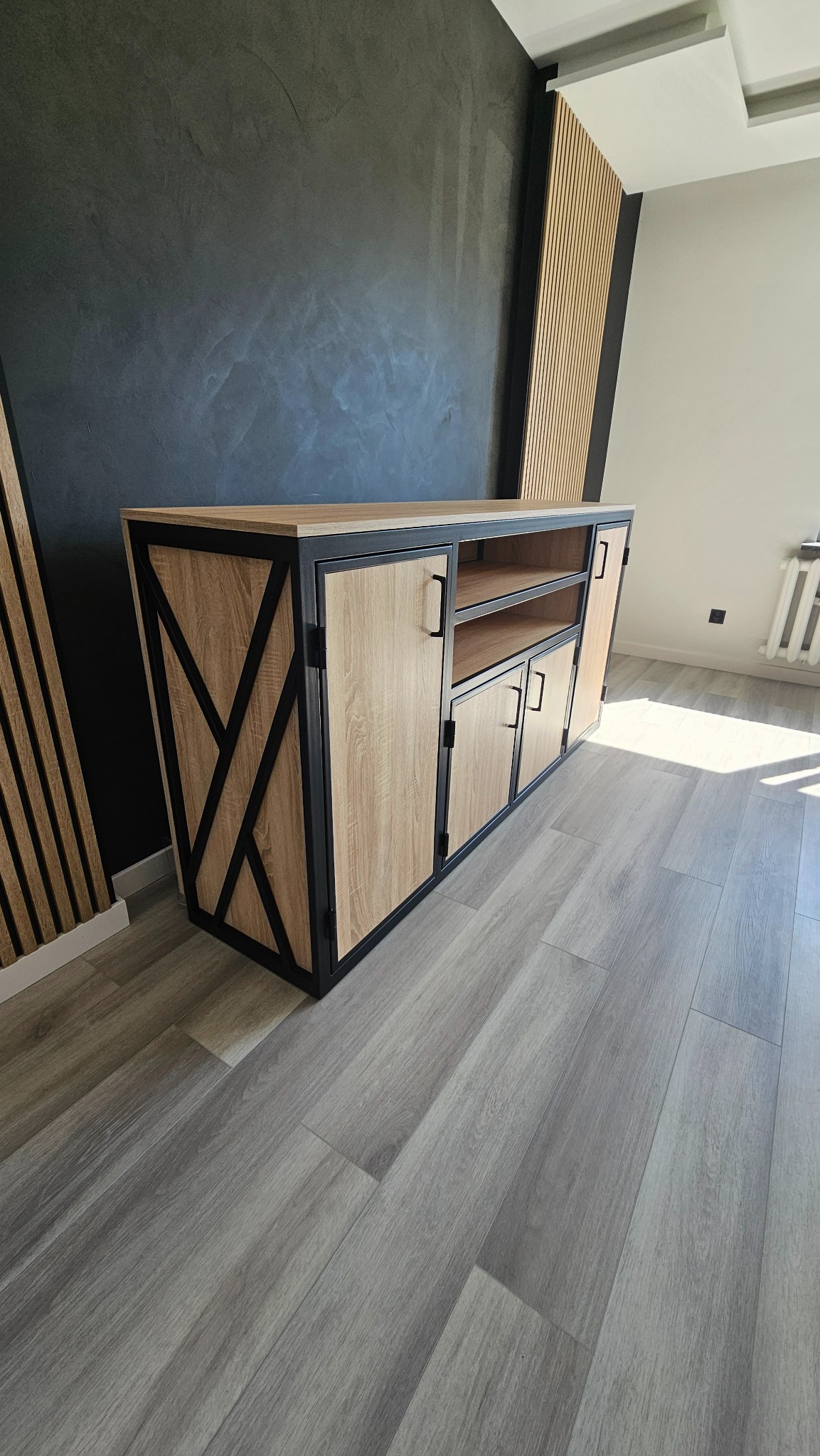 Stylowa szafka RTV z drewnianymi frontami i czarną, metalową ramą w nowoczesnym wnętrzu z szarą podłogą i czarną ścianą z dekoracyjnymi panelami.