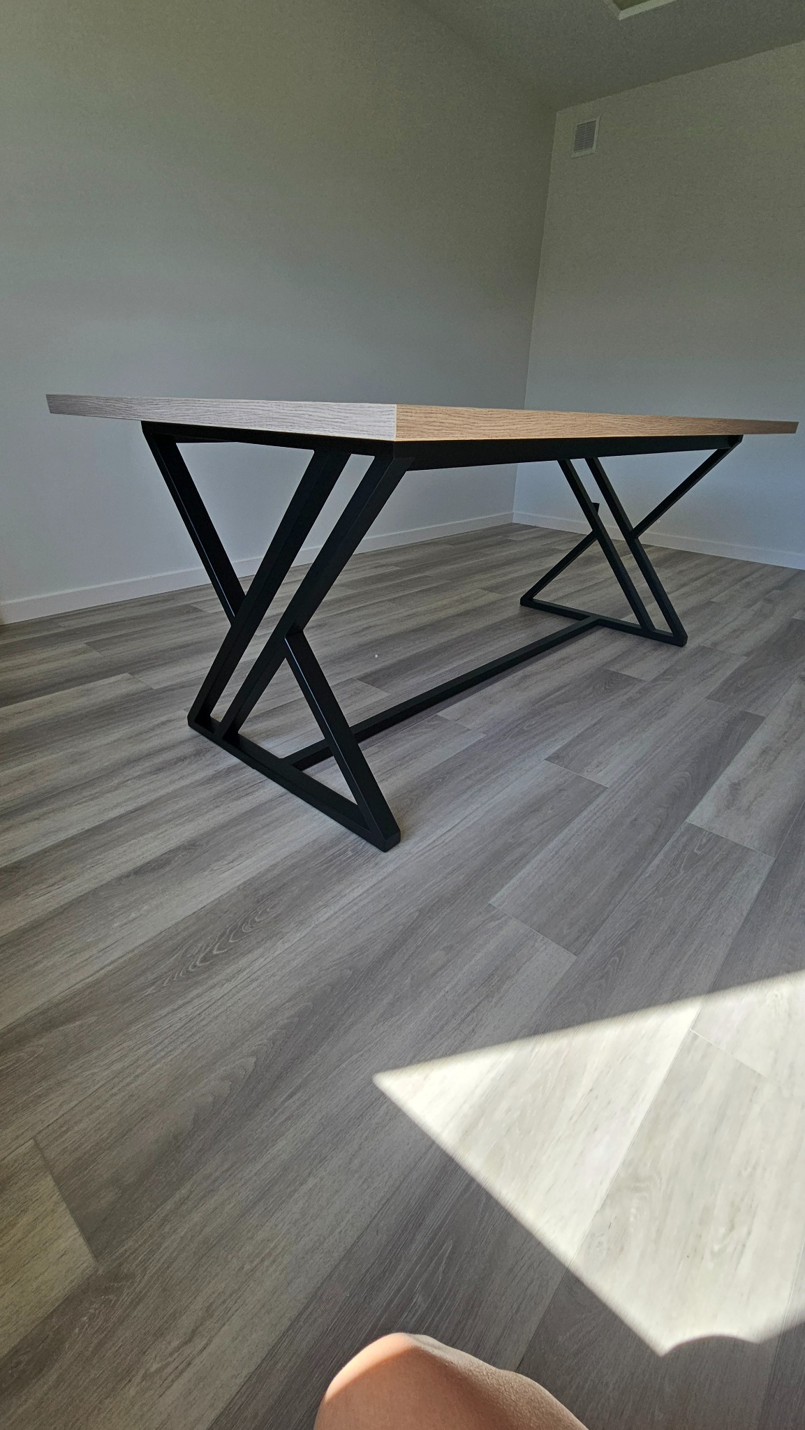 Stół z blatem w odcieniach drewna i czarną, metalową podstawą o geometrycznym kształcie. Widoczne panele podłogowe i fragment jasnej ściany.