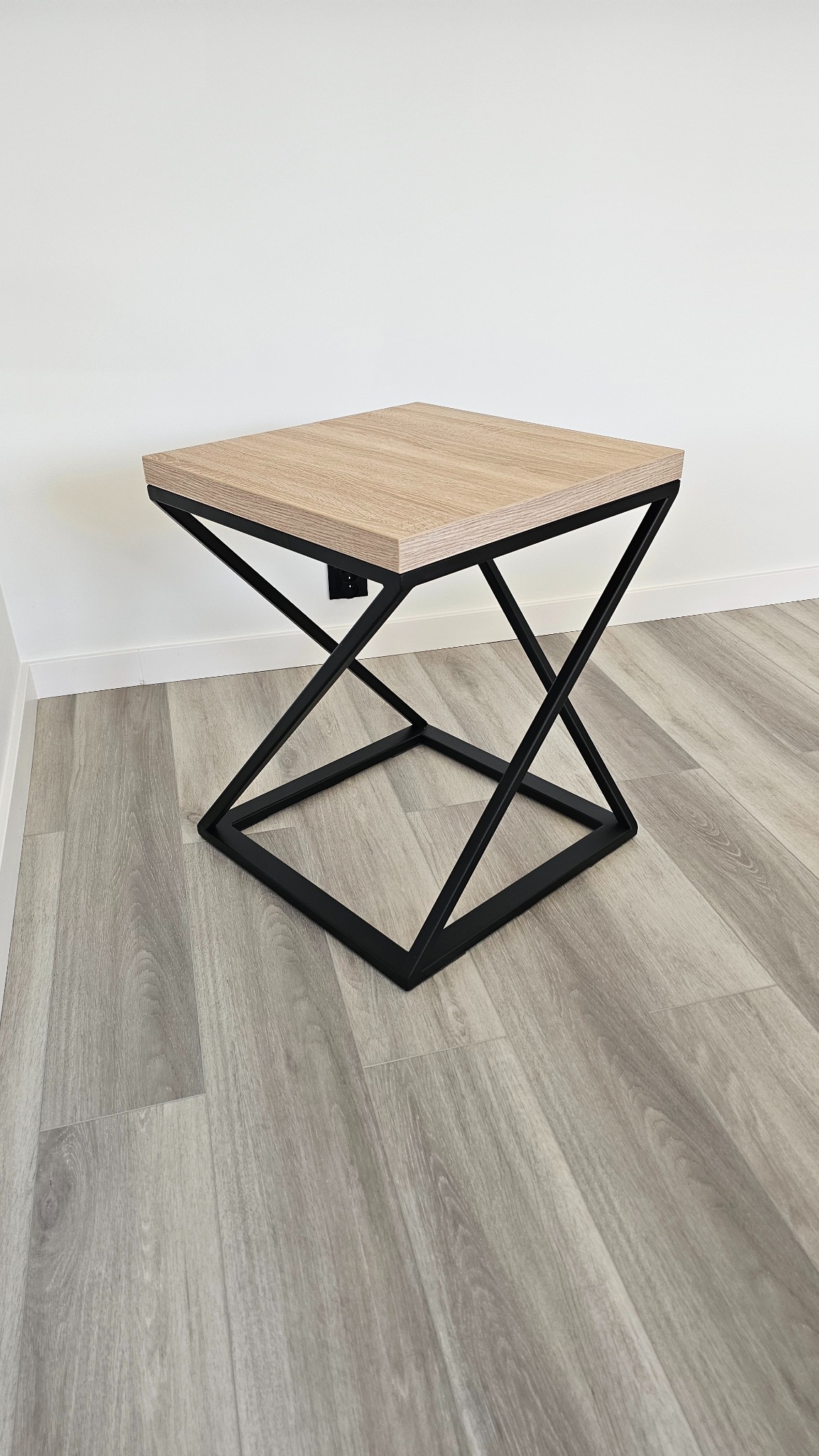 Nowoczesny stolik kawowy z blatem imitującym jasne drewno i czarną, geometryczną podstawą z metalu, na tle białej ściany i podłogi z paneli.