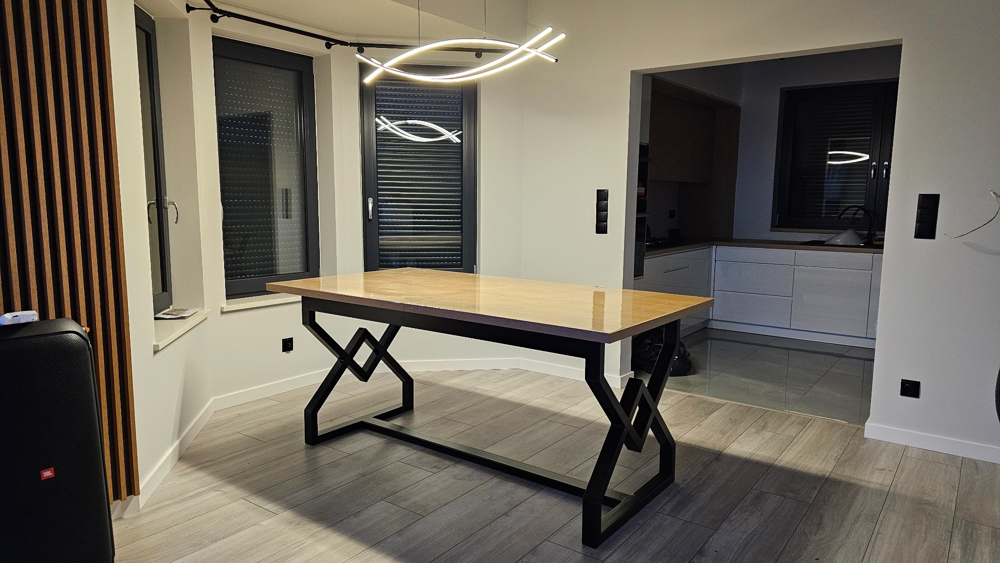 Stół z drewnianym blatem i geometryczną, metalową podstawą w nowoczesnym wnętrzu z drewnianymi lamelami i oknami z żaluzjami. Nad stołem designerska lampa.