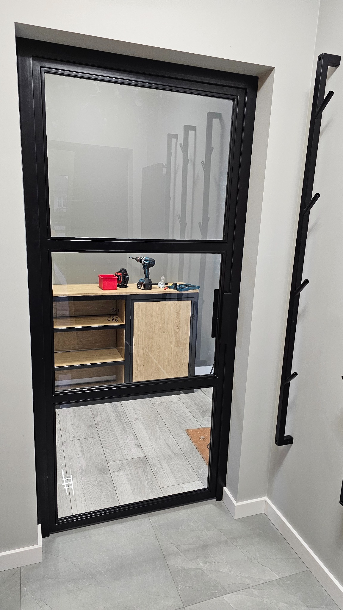 Loftowe drzwi wewnętrzne z czarną ramą i trzema szybami, widok na wnętrze z półkami i narzędziami, minimalistyczny design.