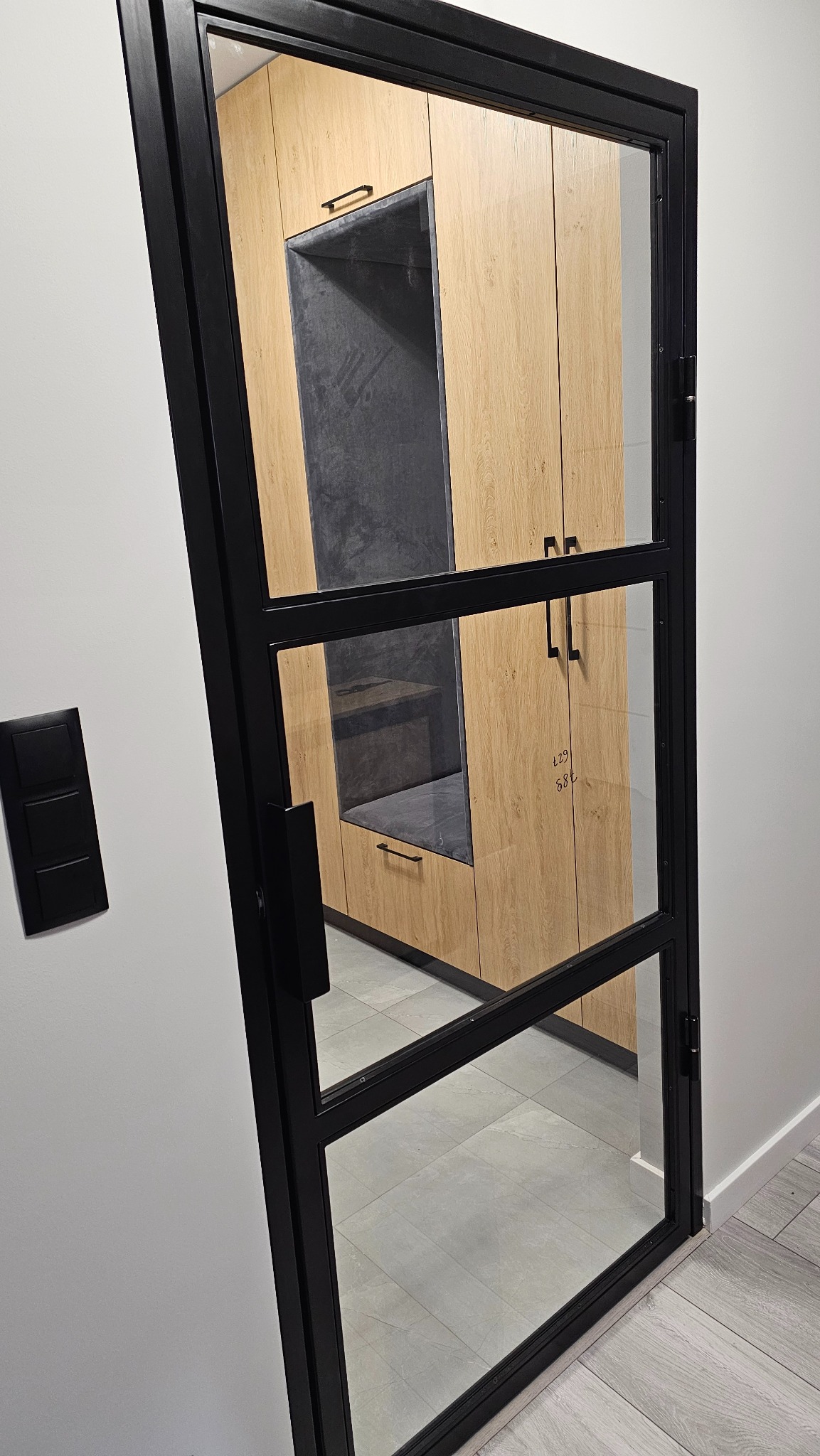 Czarna, przeszklona ścianka działowa z widokiem na zabudowę meblową z siedziskiem. Minimalistyczny design, jasne drewno, szare siedzisko, nowoczesne wnętrze.