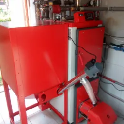 Czerwony piec z podajnikiem na paliwo stałe, widoczny panel sterowania, instalacja hydrauliczna z manometrami i zaworami, całość w pomieszczeniu z białymi płytkami.